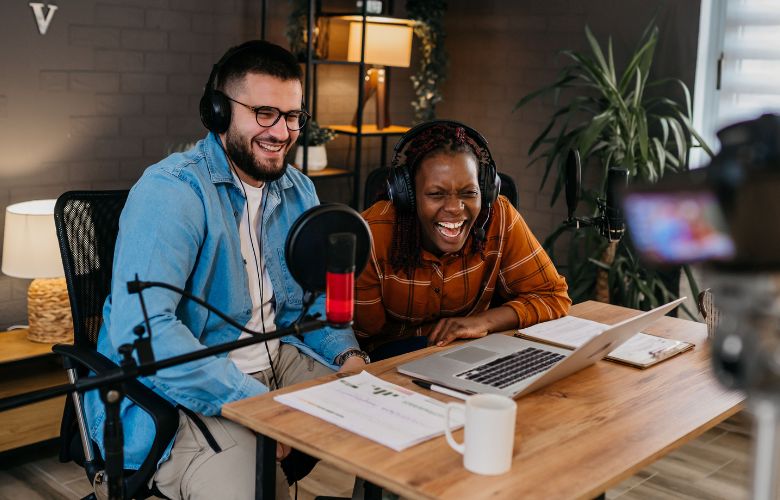 A podcast host and guest sat together recording their show. The guest is bent forwards laughing into the camera. The host is laughing along too.