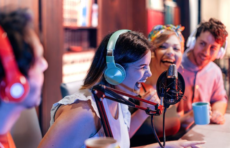 A group of four young people recording a podcast show. The host (a young lady) is roaring with laughter as her friends look on, smiling and laughing also. 