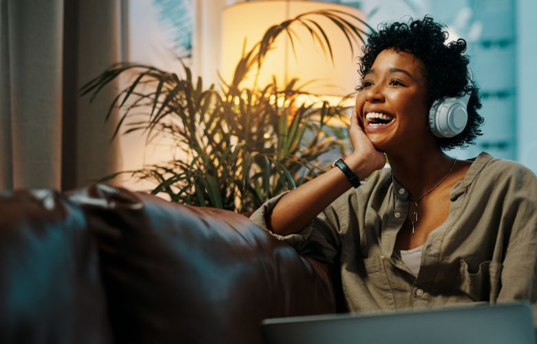 Young lady sat on a sofa smiling and laughing as she listens to a podcast on headphones.